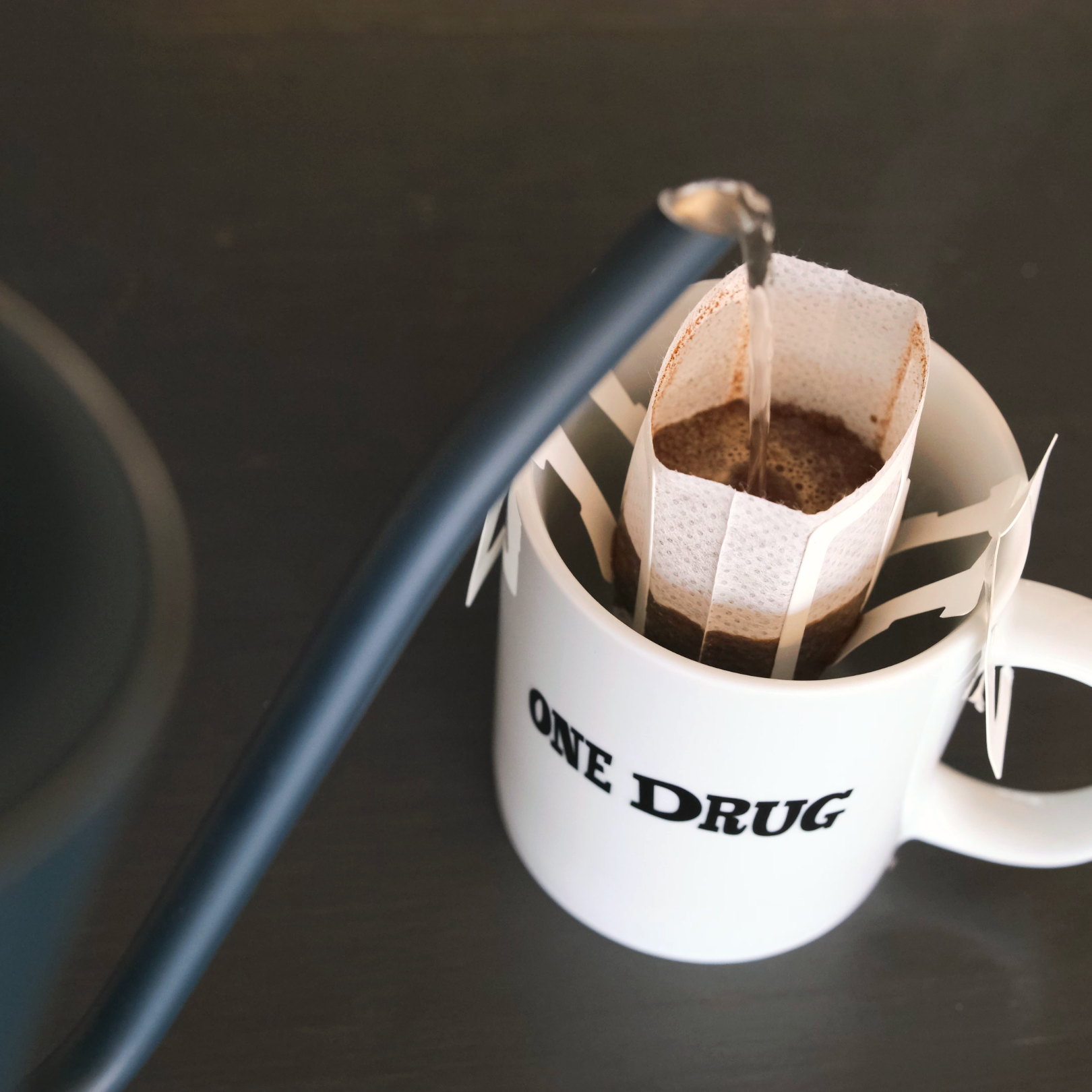 A Caffenation drip bag brewing into a One Drug mug on a dark counter